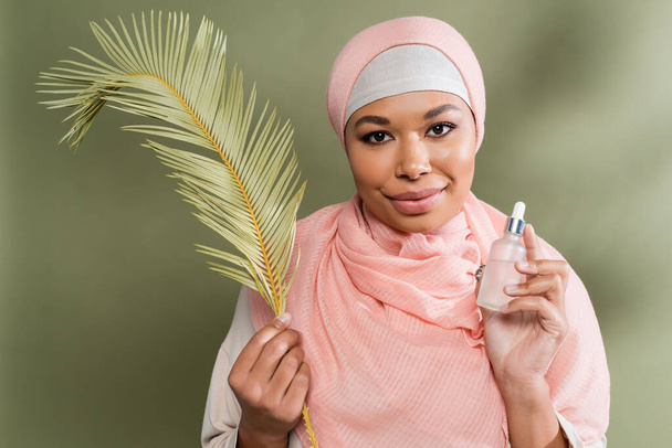 junge multirassische Frau im rosa Hijab mit kosmetischem Serum und exotischem Blatt, während sie in die Kamera auf grünem Hintergrund blickt - Foto, Bild