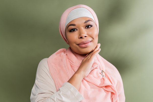 zadowolony wielorasowy muzułmanin kobieta w różowy hidżab dotykając idealny twarz i patrząc na aparat na zielonym tle - Zdjęcie, obraz
