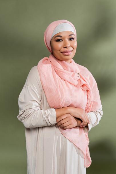mujer multirracial feliz en hijab rosa y vestido abaya tradicional sonriendo a la cámara sobre fondo verde - Foto, Imagen