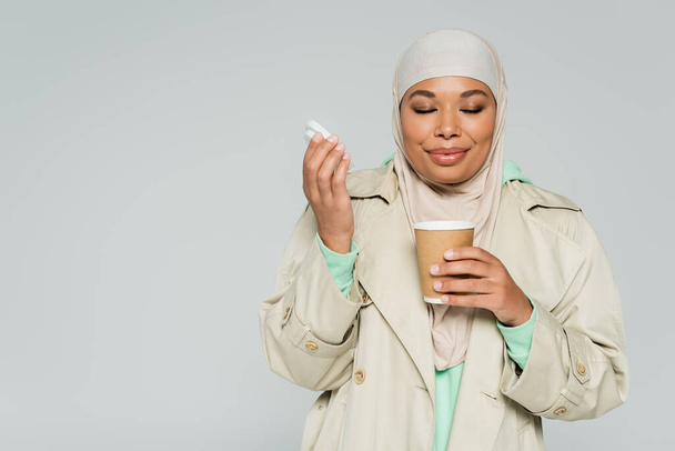 jovem mulher multirracial em casaco de trincheira e hijab desfrutando de sabor de café aromático em xícara de papel isolado em cinza - Foto, Imagem