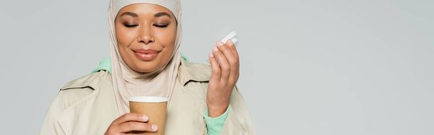 mujer musulmana multirracial complacido con los ojos cerrados disfrutando del sabor del café en taza de papel aislado en gris, pancarta - Foto, imagen