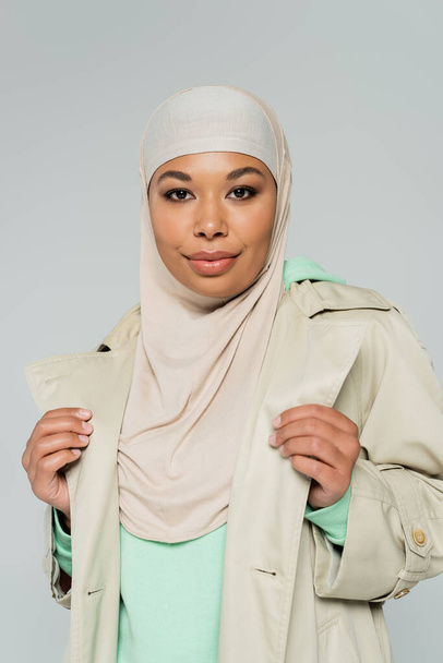 portrait of stylish multiracial woman in hijab and trench coat looking at camera isolated on grey - Zdjęcie, obraz