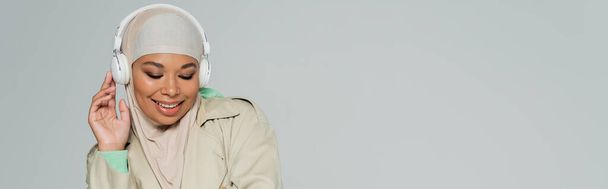 mulher muçulmana multirracial elegante e feliz com olhos fechados ouvindo música em fones de ouvido sem fio isolados em cinza, banner - Foto, Imagem