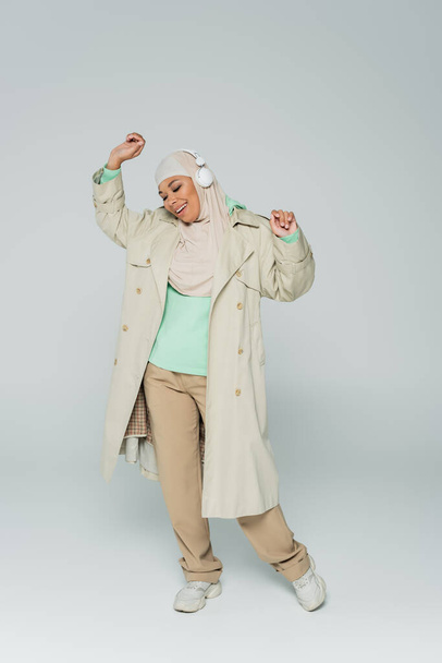 full length of carefree and fashionable multiracial muslim woman listening music in headphones and dancing on grey  - Fotoğraf, Görsel