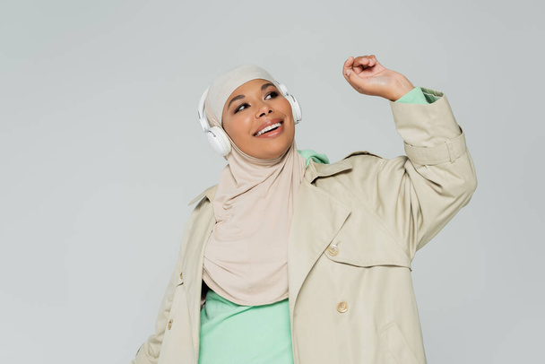 přešťastný multiracial žena v trench kabát a hidžáb poslech hudby v bezdrátových sluchátek izolovaných na šedé - Fotografie, Obrázek