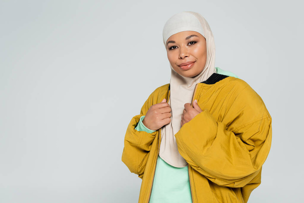muslim multiracial žena s úsměvem na kameru při pózování v hidžábu a žluté bombardér bunda izolované na šedé - Fotografie, Obrázek