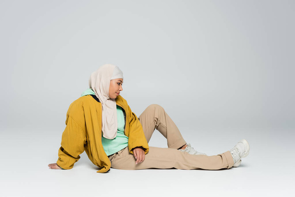 full length of multiracial woman in hijab and yellow jacket with beige pants sitting on grey background - Photo, Image