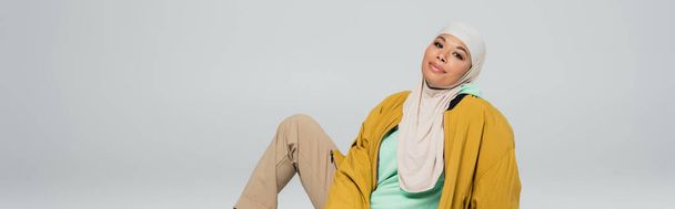 trendy multiracial muslim woman in hijab and yellow jacket smiling at camera while sitting isolated on grey, banner - Fotó, kép