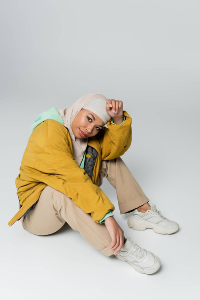 full length of stylish multiracial woman in hijab and yellow jacket sitting and looking at camera on grey background - Фото, зображення