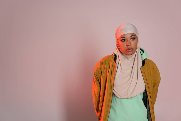 thoughtful multiracial woman in yellow jacket and muslim hijab looking away on pinkish grey background - Photo, Image