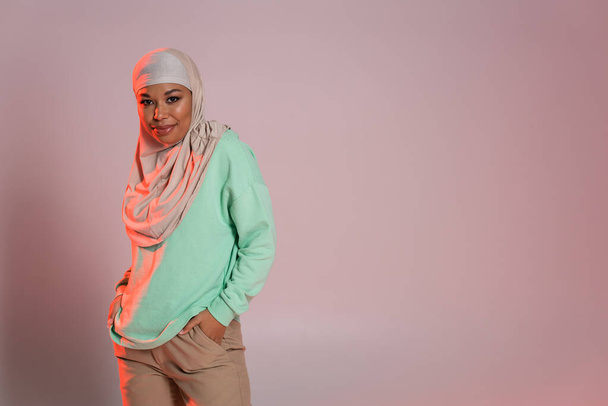 smiling multiracial woman in hijab and green long sleeve shirt posing with hands in pockets of beige pants on pinkish grey background - Photo, Image