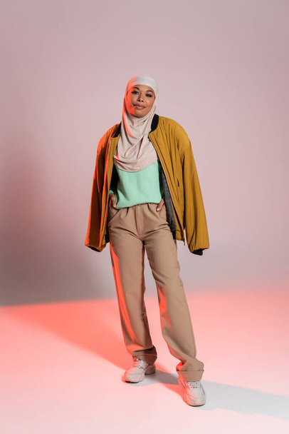 full length of multiracial muslim woman in yellow jacket and beige pants with sneakers on grey and pink background - 写真・画像