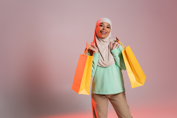 joyful multiracial woman in trendy casual attire and traditional hijab holding yellow shopping bags on pinkish grey background - Photo, Image
