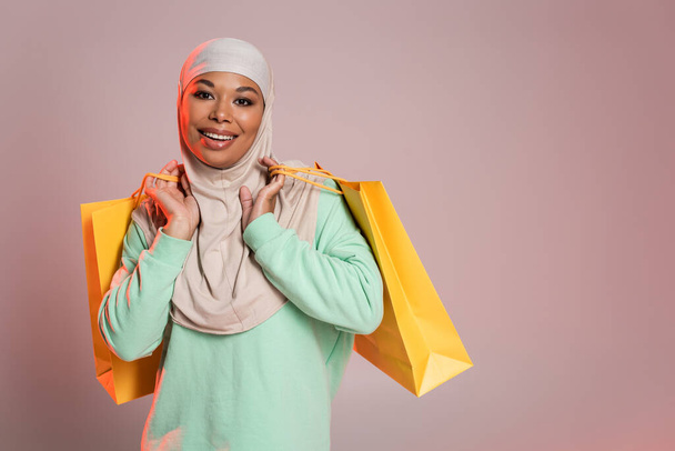 nadšené multiraciální žena v muslimském hidžábu drží žluté nákupní tašky a usmívá se na kameru na narůžovělé šedé - Fotografie, Obrázek
