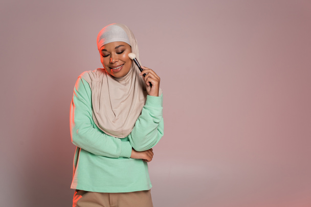 pleased multiracial muslim woman in hijab holding cosmetic brush and smiling with closed eyes on pinkish grey background - Photo, Image