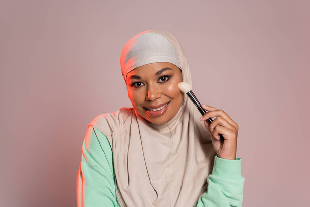 vidám multiracionális nő hidzsáb és hosszú ujjú ing kezében kozmetikai kefe és mosolygott a kamera rózsaszín és szürke - Fotó, kép