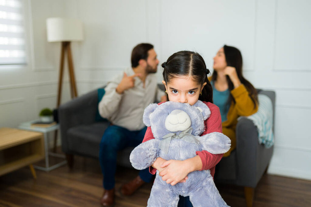 Adorabile bambina che abbraccia un orsacchiotto spaventato e triste mentre ascolta i suoi genitori litigare in salotto - Foto, immagini