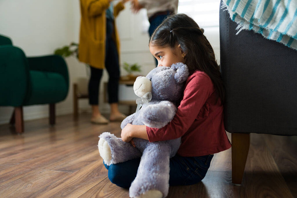 Λυπημένο παιδί που κάθεται μόνο του αγκαλιάζοντας ένα αρκουδάκι ενώ δείχνει τρομαγμένο που η μαμά και ο μπαμπάς ουρλιάζουν και τσακώνονται στο σπίτι - Φωτογραφία, εικόνα