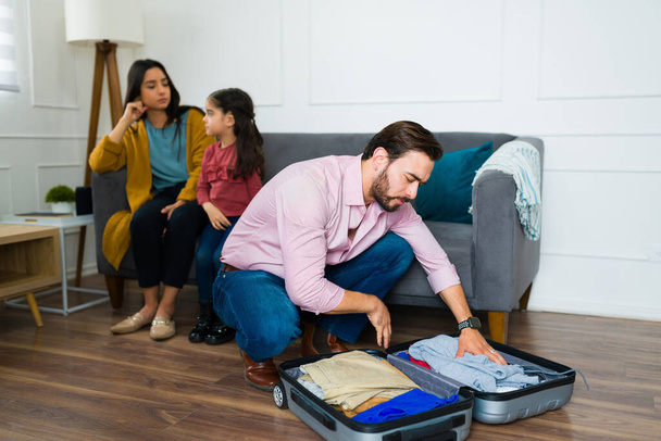 Upset caucasian man packing his suitcase after divorce and leaving his family because of their bad marriage problems  - 写真・画像