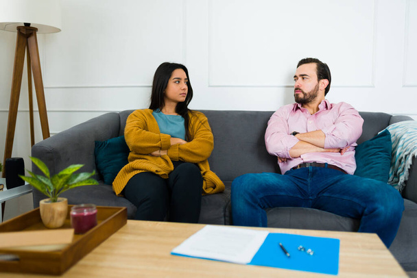 Dühös fiatal nő és férfi szomorúnak tűnnek, keresztbe tett karokkal ülnek a kanapén, és azon gondolkodnak, hogy aláírják a válási papírokat. - Fotó, kép