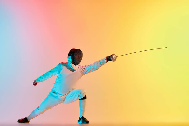 Long attack. Young man, male fencer with sword practicing in fencing over gradient pink-yellow background in neon light. Sportsman shows fencing technique. Copyspace for ad. - Foto, imagen
