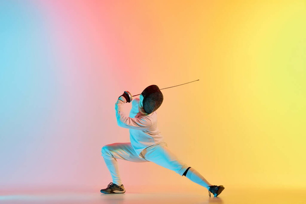 Fencer takes a stand. Young man, male fencer with sword practicing in fencing over gradient pink-yellow background in neon light. Sportsman shows fencing technique. Copyspace for ad. - Foto, Bild