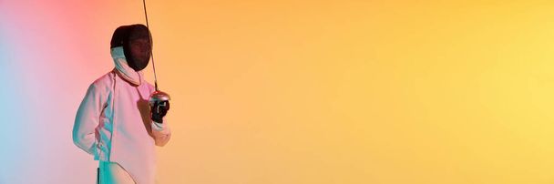 Hombre joven, esgrimista masculino en traje de esgrima y máscara de casco de pie con espada sobre el degradado fondo rosa-amarillo en luz de neón. Banner horizontal con copyspace para anuncio, texto. - Foto, imagen