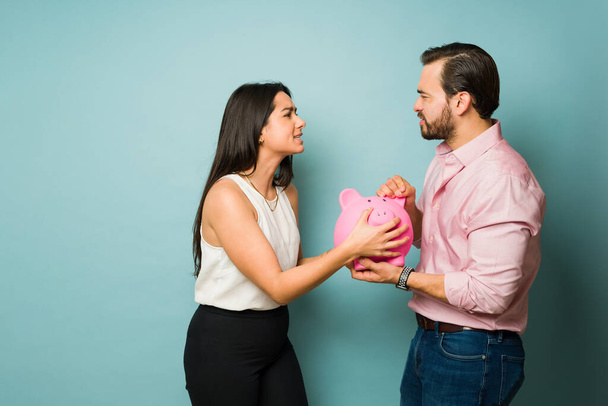 Латиноамериканка и кавказский женатый мужчина держат копилку и дерутся из-за денег или их финансов - Фото, изображение