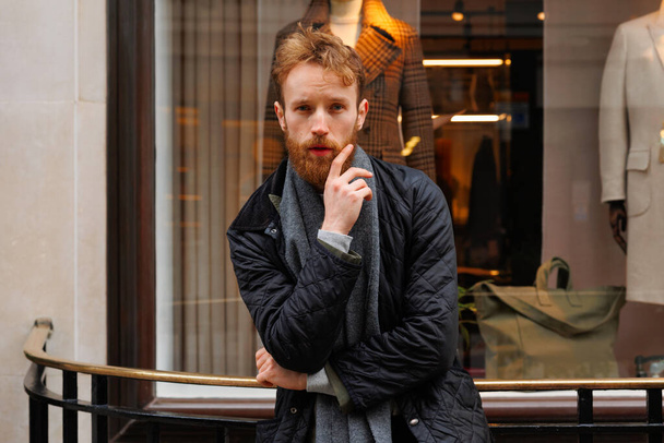 Bearded man in jacket and scarf posing outdoors near clothing store - 写真・画像
