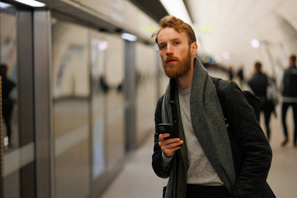 Bearded man is standing on the platform of a subway station with a smartphone in his hands, looking at the train - Foto, Bild