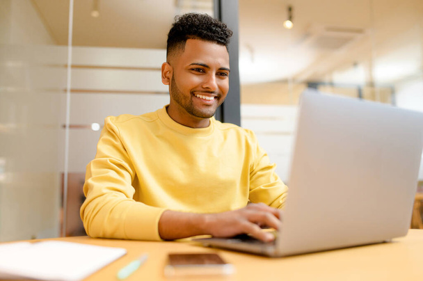 Successful freelancer. Happy millennial Indian man sitting at desk with laptop, smiling and typing, enjoying remote work, male student studying on the distance - 写真・画像