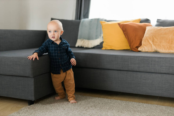 Petit garçon riant s'amusant debout près du canapé dans le salon à la maison. Adorable bébé faisant les premiers pas seul. Bonne enfance et garde d'enfants - Photo, image