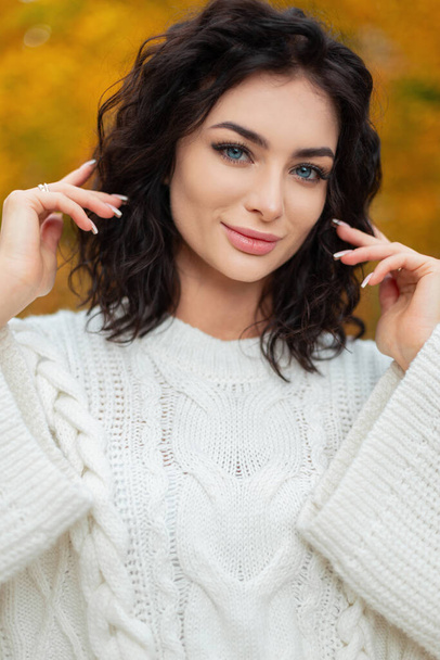 Ευτυχισμένη όμορφη γυναίκα με καταπληκτικά μπλε μάτια και σγουρά μαλλιά με μοντέρνο τρόπο πλεκτό λευκό πουλόβερ στέκεται σε ένα φθινοπωρινό κίτρινο πάρκο και κοιτάζει την κάμερα - Φωτογραφία, εικόνα