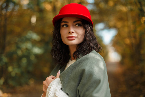 Élégante belle jeune jolie femme avec un chapeau rouge bouclé et un pull blanc tricoté à la mode avec une écharpe verte se promène dans le parc d'automne - Photo, image