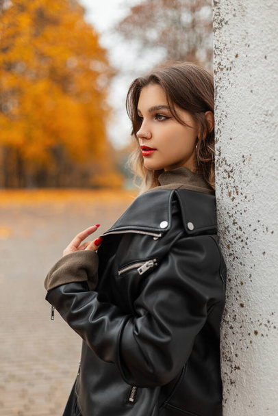 Modieus mooi meisje met rode lippen in een mode zwart leren jas staat in de buurt van een witte muur in een herfst park - Foto, afbeelding