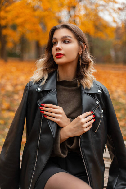 Şık, güzel, kırmızı dudaklı, siyah deri ceketli ve yeşil kazaklı bir genç kız sonbahar parkında parlak sarı yapraklarıyla oturuyor. - Fotoğraf, Görsel