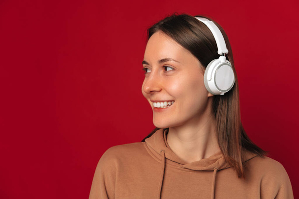 Pretty smiling woman is wearing headphones and looking aside at copy space over red background. - Foto, Bild