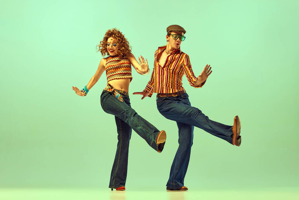 Динамичный портрет счастливого мужчины и женщины в ретро-цветных рубашках и раздутых джинсах, танцующих энергичный танец на зеленом фоне. Концепция модных тенденций 70-х, 80-х годов, музыки, образа жизни хиппи - Фото, изображение
