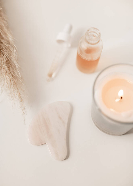 From above of face serum placed on table with pink Gua Sha tool and burning candle for spa procedure in beauty salon - Zdjęcie, obraz