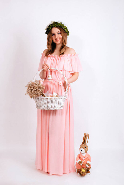 A girl in a pink dress and a wreath on her head is holding Easter basket with eggs on a white background. Smiling young woman is holding Easter basket with eggs on a white background. - Φωτογραφία, εικόνα