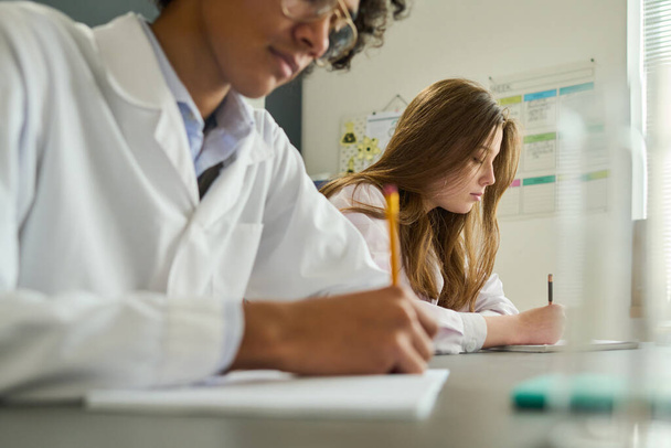 Menina jovem no labcoat fazendo anotações no livro de anotações durante o trabalho de laboratório na lição de química enquanto se senta ao lado de seu colega de classe - Foto, Imagem