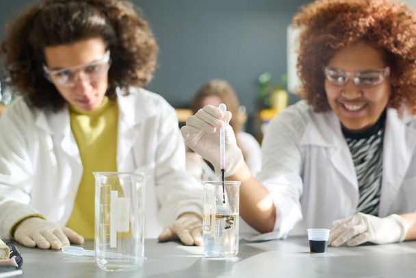 Teini-ikäinen naisopiskelija käsineissä, silmälaseissa ja labcoat pudottamalla mustaa nestemäistä ainetta putkeen läpinäkyvällä nesteellä kemian tunnilla - Valokuva, kuva