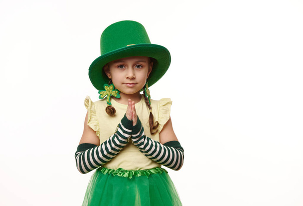 Irish lovely little girl, wearing green hat, striped gloves and clover leaf earrings, keeping her hands palms together, isolated on white background with copy space. Saint Patricks Day celebration - Photo, image