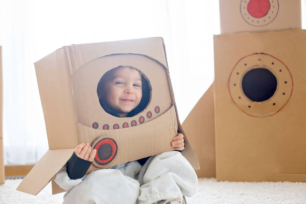 Sweet toddler boy, dressed as an astronaut, playing at home with cardboard rocket and handmade helmet from box - Foto, Imagen