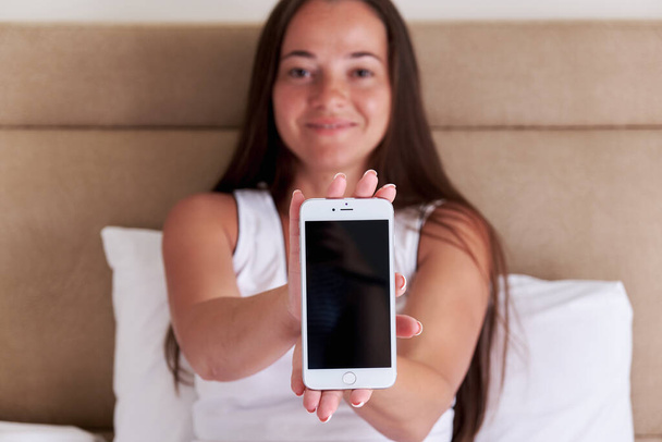 Smartphone with blank screen, mobile app mockup. Happy woman demonstrates smartphone screen while sitting in bed in the morning. Template for design. - Zdjęcie, obraz