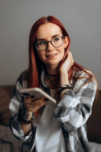 A red-haired girl with glasses and a plaid shirt is holding a phone smiling and looks away - Fotoğraf, Görsel
