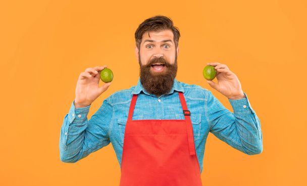 Surprised man in red apron holding fresh limes citrus fruits yellow background, fruiterer. - 写真・画像