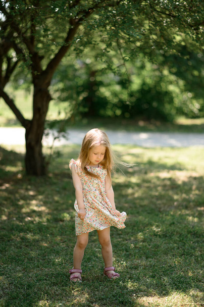 Little girl 3 years old in a summer sundress in the park. Summer time.The baby is playing with her dress. - Foto, Bild