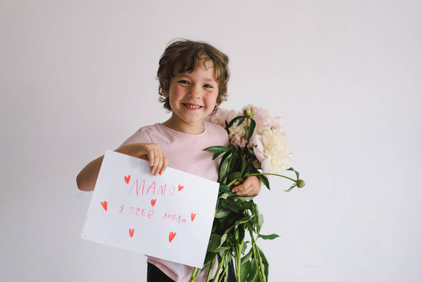 Cheerful happy child with Peonys bouquet and a card for mom in Ukrainian language. Smiling little boy on white background. Mothers Day concept - Photo, Image