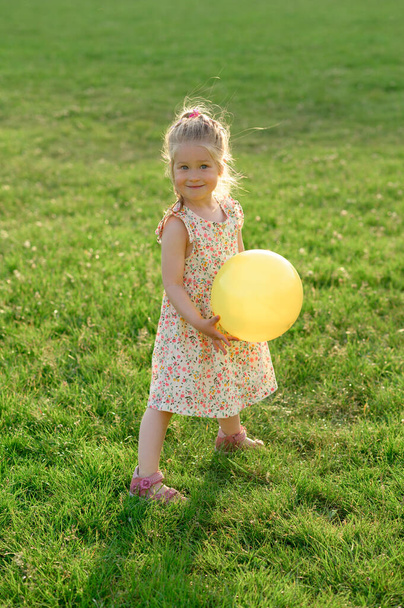 Elinde sarı bir topla, açık bir alanda, yaz elbisesi içinde üç yaşında küçük bir kız. Kız mutlu.. - Fotoğraf, Görsel